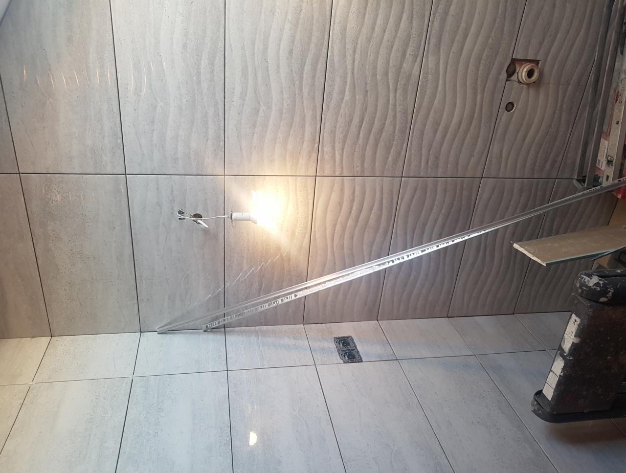 Ściana i podłoga wyłożone szarymi płytkami z delikatnym wzorem falistym, widoczne narzędzie do cięcia płytek, gniazdko elektryczne bez osprzętu oraz żarówka tymczasowa.