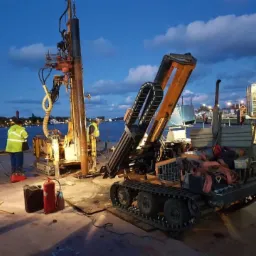 Prace geologiczne na nabrzeżu: pomarańczowa wiertnica gąsienicowa w trakcie wiercenia, dwóch pracowników w odblaskowych kamizelkach i sprzęt oświetleniowy przy akwenie wodnym pod wieczornym niebem.