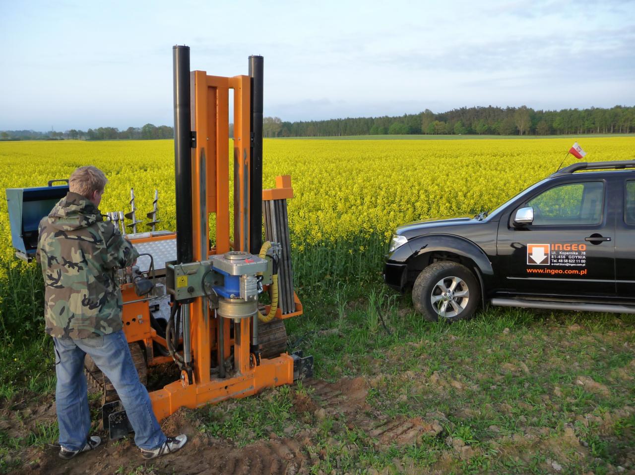 Pracownik obsługuje wiertnicę na gąsienicach na tle pola rzepaku i czarnego samochodu terenowego z logo firmy.