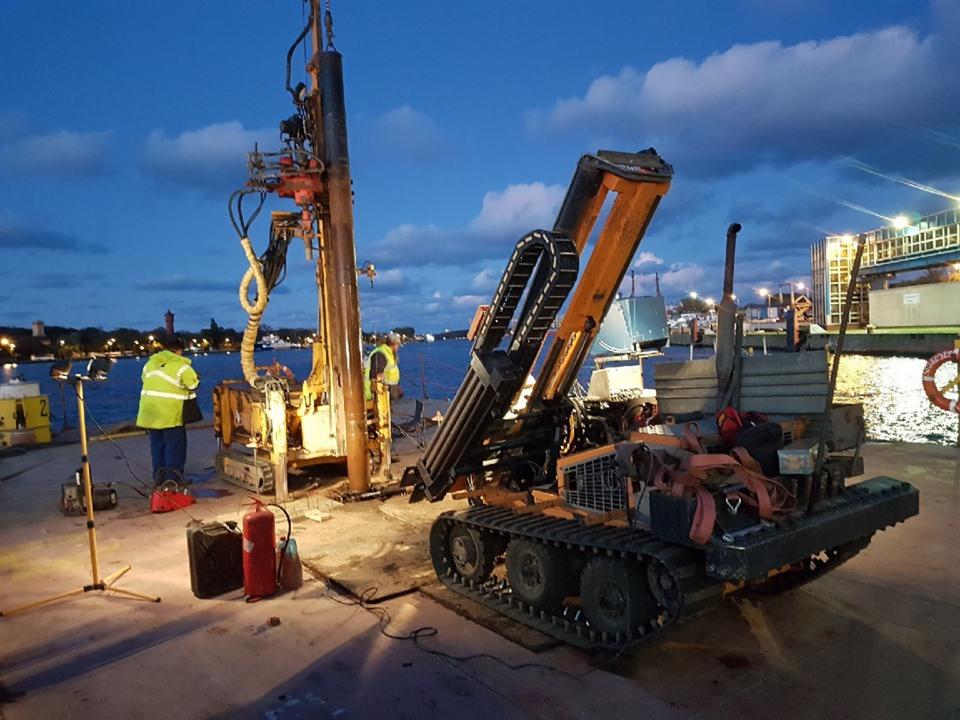 Prace geologiczne na nabrzeżu: pomarańczowa wiertnica gąsienicowa w trakcie wiercenia, dwóch pracowników w odblaskowych kamizelkach i sprzęt oświetleniowy przy akwenie wodnym pod wieczornym niebem.