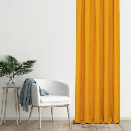 Jasny pokój z musztardową zasłoną, białym fotelem z narzuconym błękitnym pledem i stolikiem z rośliną w szklanym wazonie.