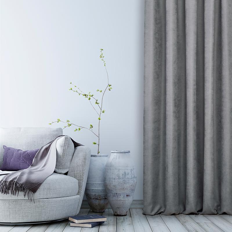 Minimalistyczny salon z szarym fotelem, fioletową poduszką, pledem, ceramicznymi wazonami z gałązkami i długą, szarą zasłoną na drewnianej podłodze.