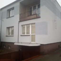 Dwukondygnacyjny dom z balkonem, pomalowany w odcieniach szarości i brązu, z dekoracyjną balustradą balkonową i metalową drabinką na ścianie bocznej.