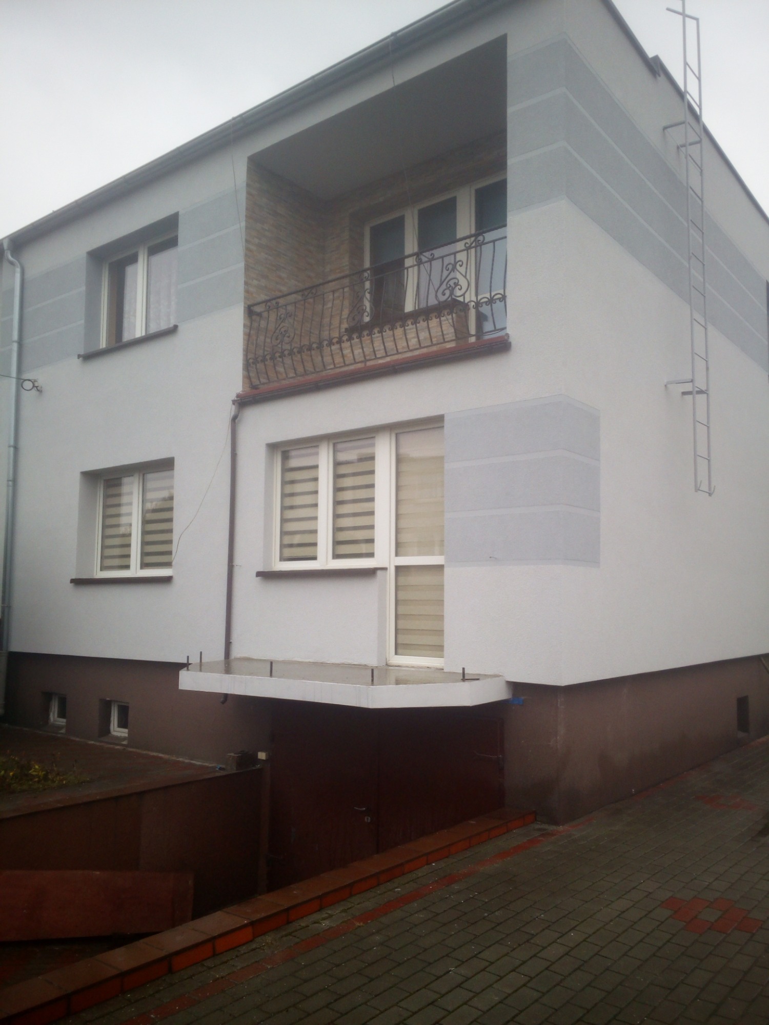 Dwukondygnacyjny dom z balkonem, pomalowany w odcieniach szarości i brązu, z dekoracyjną balustradą balkonową i metalową drabinką na ścianie bocznej.