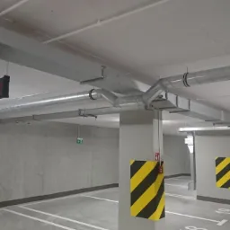 Instalacja wentylacji w garażu podziemnym: widoczne rury wentylacyjne pod sufitem, oznaczenia ostrzegawcze na filarach, tablica informacyjna.