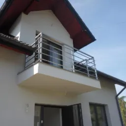 Elewacja domu z balkonem zabezpieczonym metalową balustradą o prostym, nowoczesnym designie, widok z dołu na tle błękitnego nieba.