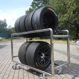 Wózek transportowy ze stali nierdzewnej z kompletem opon wyścigowych Hankook na dwóch poziomach, na kostce brukowej, z widocznym drzewem w tle.
