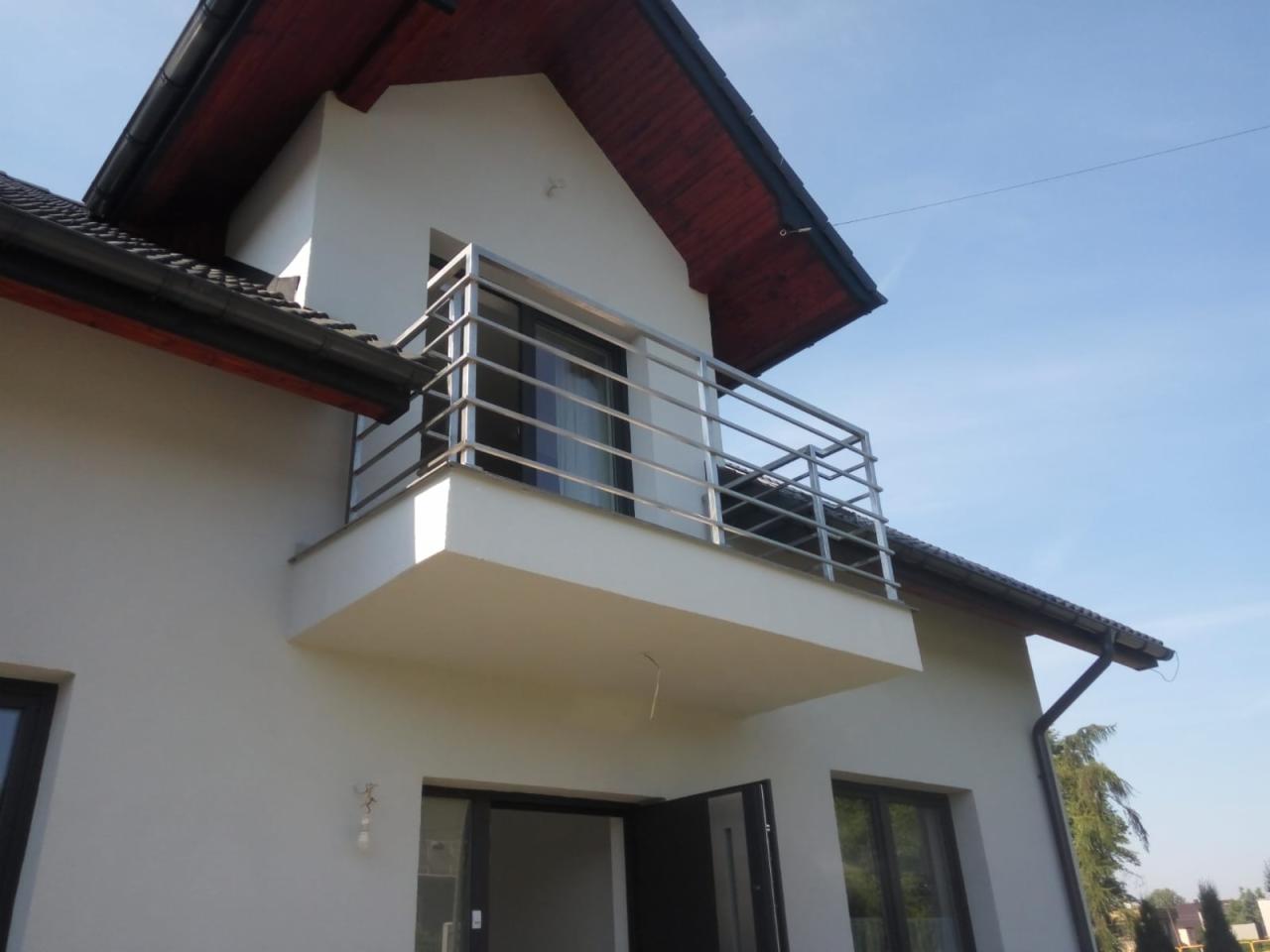 Elewacja domu z balkonem zabezpieczonym metalową balustradą o prostym, nowoczesnym designie, widok z dołu na tle błękitnego nieba.