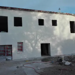 Świeżo otynkowana elewacja budynku z widocznymi otworami okiennymi i drzwiowymi, częściowo zabezpieczonymi folią, z rozstawioną drabiną i śladami prac wykończeniowych na ziemi.