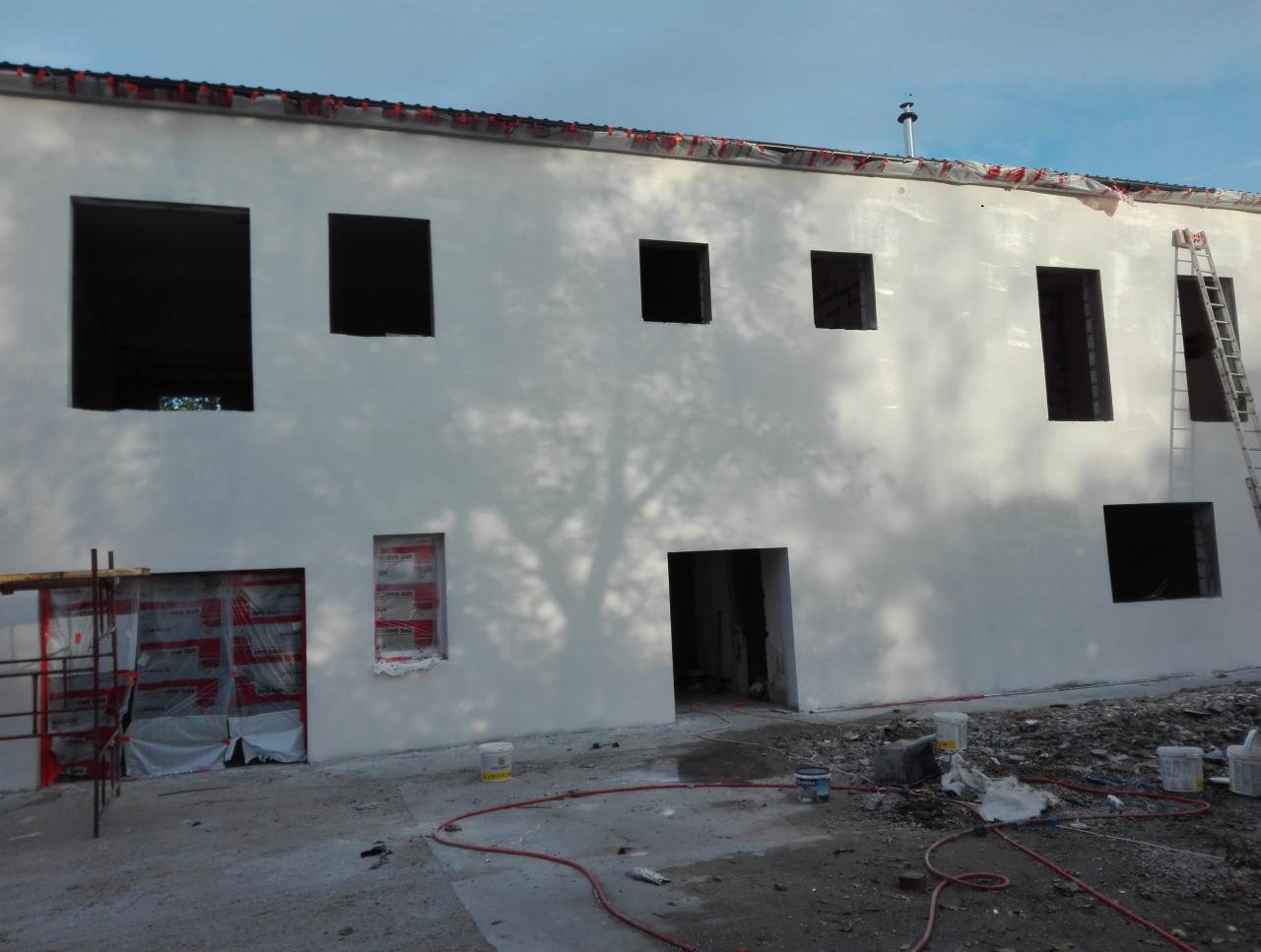 Świeżo otynkowana elewacja budynku z widocznymi otworami okiennymi i drzwiowymi, częściowo zabezpieczonymi folią, z rozstawioną drabiną i śladami prac wykończeniowych na ziemi.