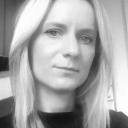 Czarno-biały portret kobiety z blond włosami, skierowanej lekko w prawo, z delikatnym uśmiechem i neutralnym wyrazem twarzy. Ujęcie od przodu, z widocznym fragmentem ramion.