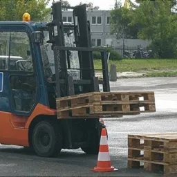 Wózek widłowy z oznaczeniem 'L' unosi palety, na placu manewrowym widoczne pachołki.