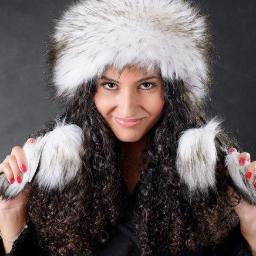 Avito - Uśmiechnięta kobieta z kręconymi włosami prezentuje futrzaną czapkę z pomponem i nausznikami, ubrana w czarny płaszcz.