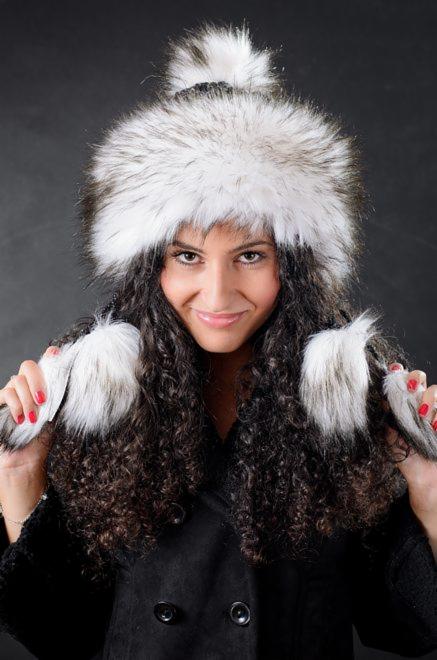 Uśmiechnięta kobieta z kręconymi włosami prezentuje futrzaną czapkę z pomponem i nausznikami, ubrana w czarny płaszcz.