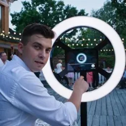 Mężczyzna w białej koszuli pozuje przy fotobudce w kształcie pierścienia z oświetleniem LED na tle imprezy plenerowej z girlandami świetlnymi.