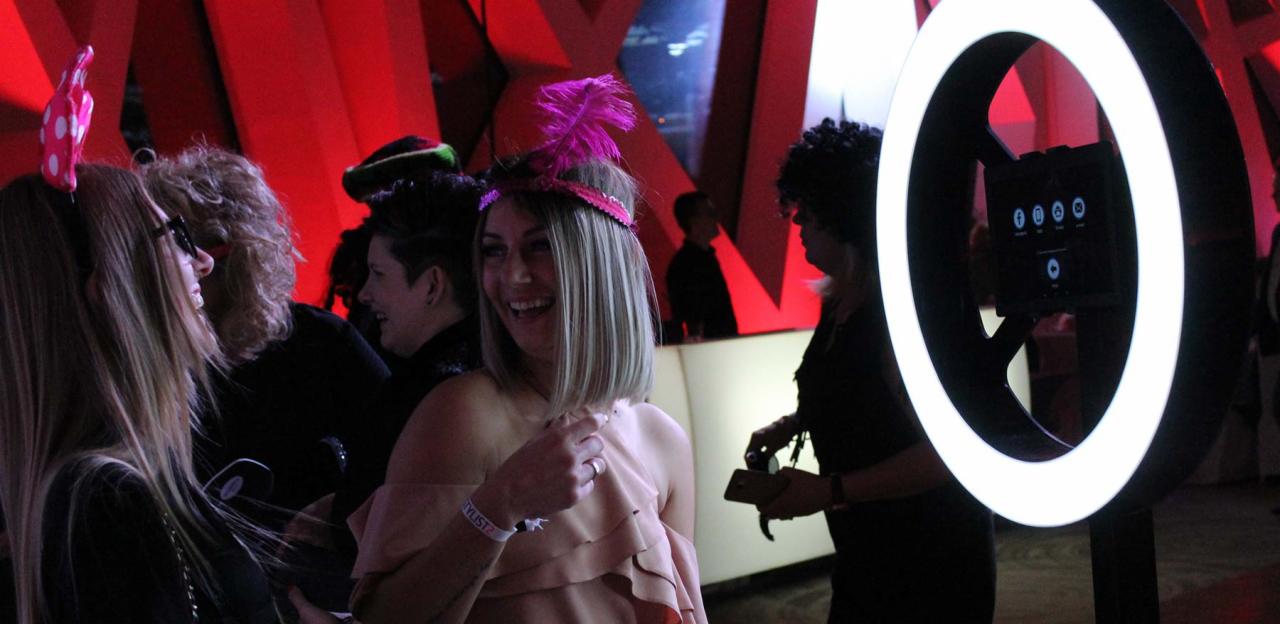 Uśmiechnięta kobieta w różowej sukience i opasce z piórami pozuje do zdjęcia w fotobudce z pierścieniowym oświetleniem. W tle widoczni inni uczestnicy imprezy i czerwone dekoracje.
