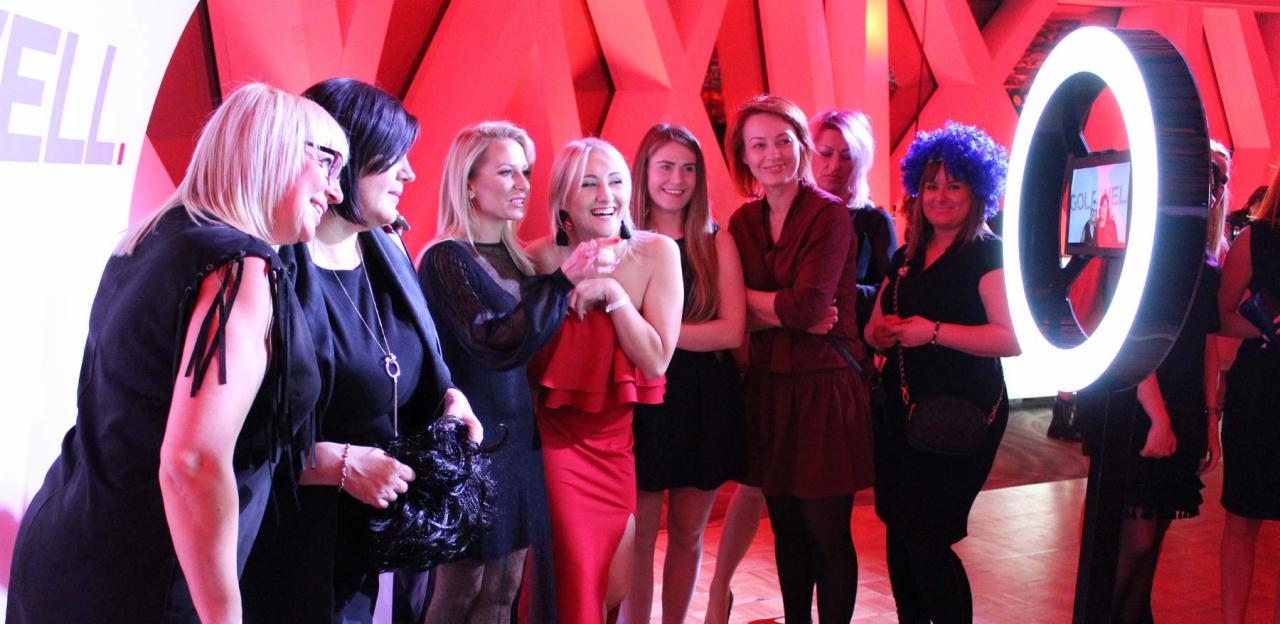 Grupa kobiet pozuje do zdjęcia przy podświetlanym okręgu fotobudki na tle czerwonej ściany z geometrycznym wzorem.