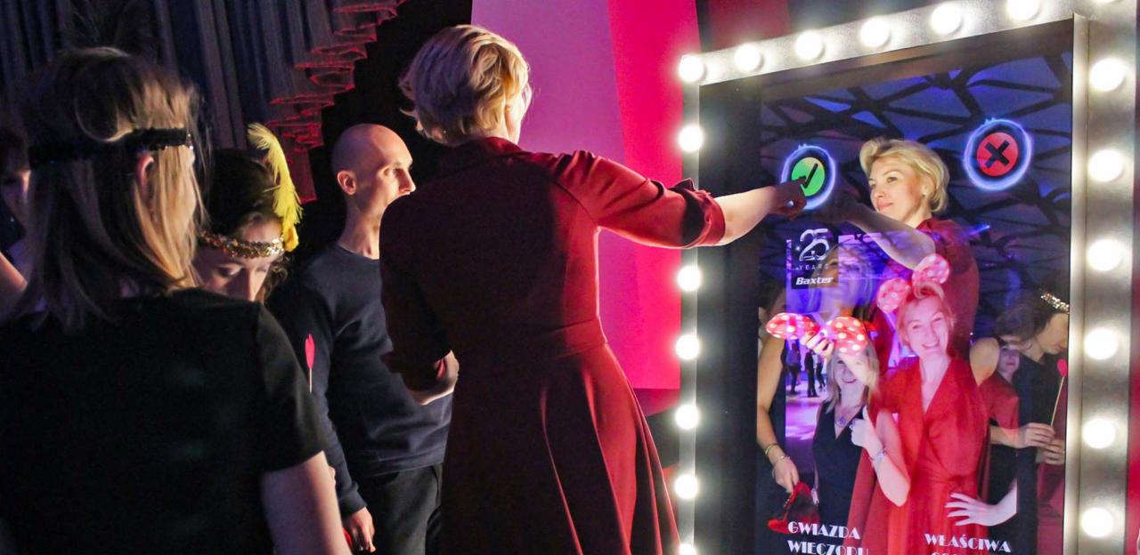 Grupa osób korzystająca z interaktywnej fotobudki z oświetleniem LED, która wyświetla wirtualne akcesoria i efekty specjalne. Kobieta w czerwonej sukience dotyka ekranu fotobudki, na którym...