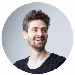 Portret uśmiechniętego mężczyzny z krótką brodą i kręconymi włosami, ubrany w czarną koszulkę, na jasnoszarym tle w okrągłej ramce.