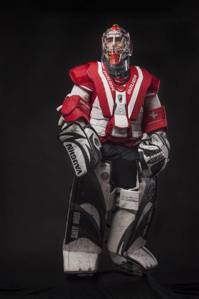 Hokejowy bramkarz w pełnym rynsztunku, z maską ozdobioną płomieniami, w pozycji gotowej do obrony krążka na czarnym tle.