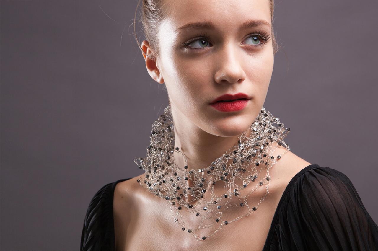 Portret kobiety z błękitnymi oczami i czerwonymi ustami, noszącej srebrny naszyjnik z czarnymi koralikami na tle szarego tła.