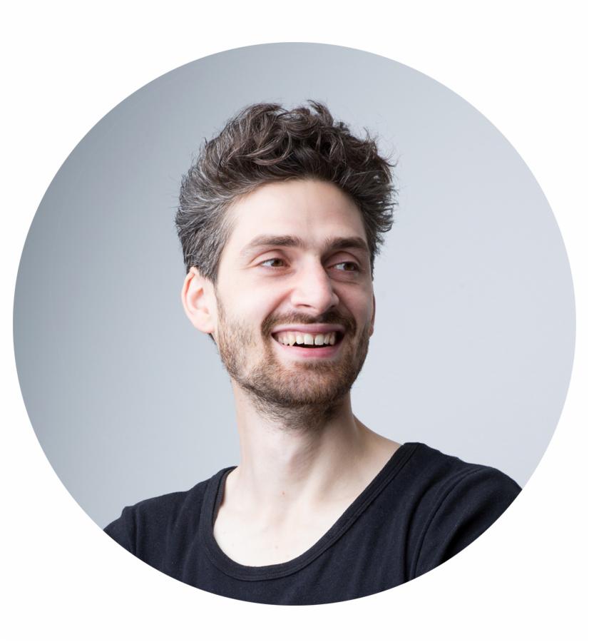 Portret uśmiechniętego mężczyzny z krótką brodą i kręconymi włosami, ubrany w czarną koszulkę, na jasnoszarym tle w okrągłej ramce.