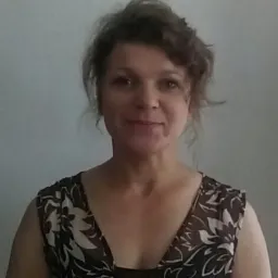 Portret kobiety o brązowych kręconych włosach, ubranej w sukienkę w kwiaty, na jasnym tle. Uśmiechnięta twarz, naturalny makijaż.