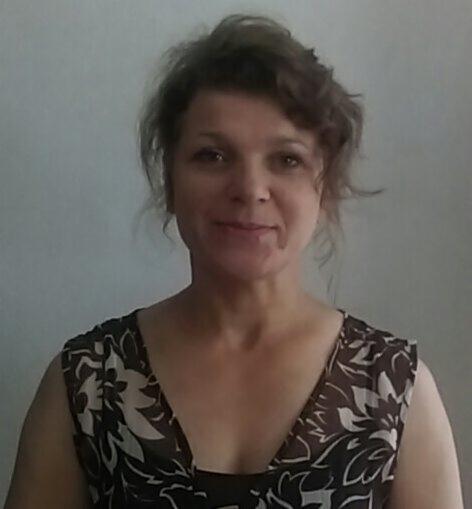 Portret kobiety o brązowych kręconych włosach, ubranej w sukienkę w kwiaty, na jasnym tle. Uśmiechnięta twarz, naturalny makijaż.