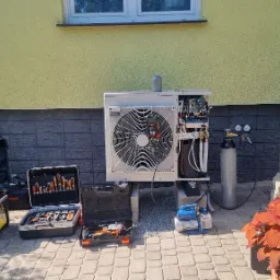 Instalacja pompy ciepła: jednostka zewnętrzna na betonowych blokach, otwarte skrzynki z narzędziami, butla z gazem i manometry, pompa próżniowa, miernik podłączony do jednostki, zieleń w tle.