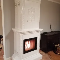 Biały, kaflowy kominek z paleniskiem, ozdobiony reliefami, na tle szarej ściany i drewnianej podłogi. W palenisku widoczny ogień.