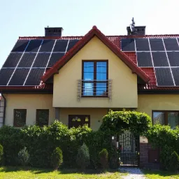 Dom jednorodzinny z panelami fotowoltaicznymi na dachu, widoczne panele na dwóch połaciach dachu, czerwona dachówka, kremowa elewacja, brama wejściowa z roślinnością.