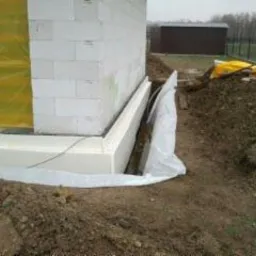 Fundament budynku w trakcie ocieplania styropianem, zabezpieczony folią, widoczny wykop i pryzma ziemi.