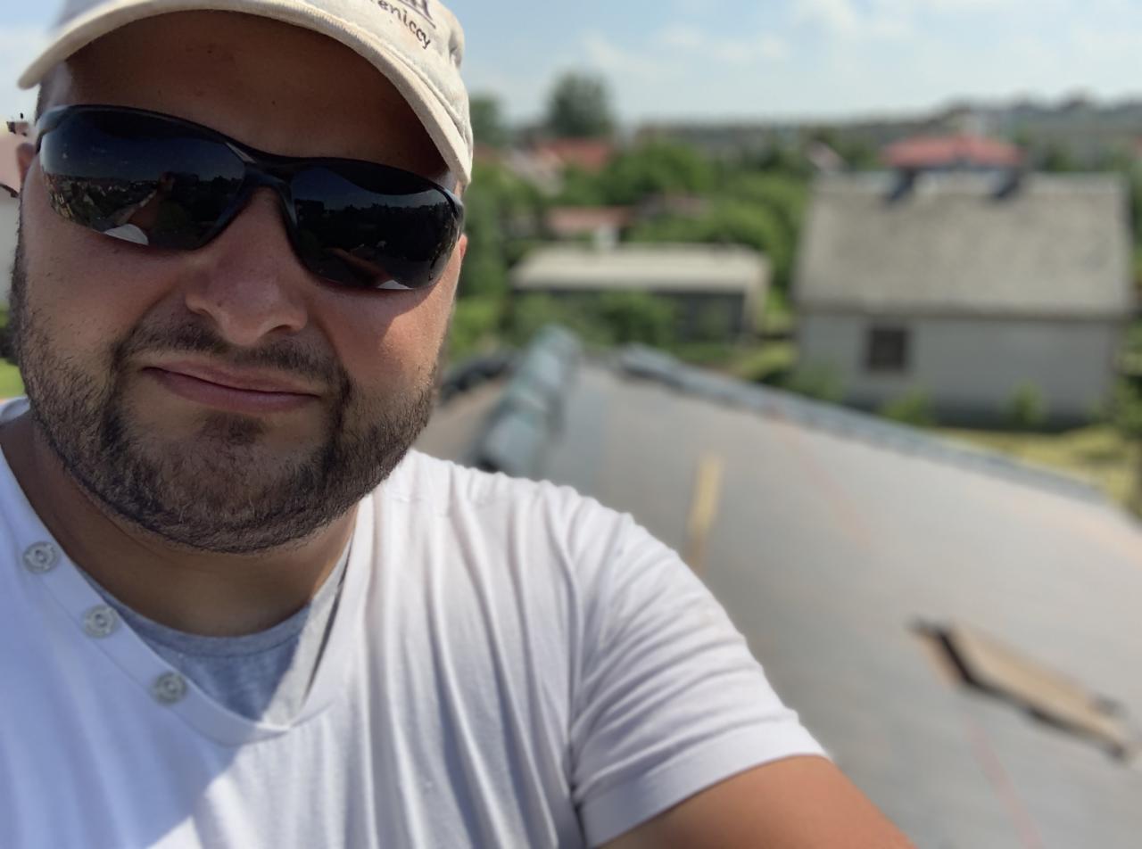 Mężczyzna w okularach na tle dachu z widocznymi uszkodzeniami i budynkami w oddali, wykonujący autoportret.