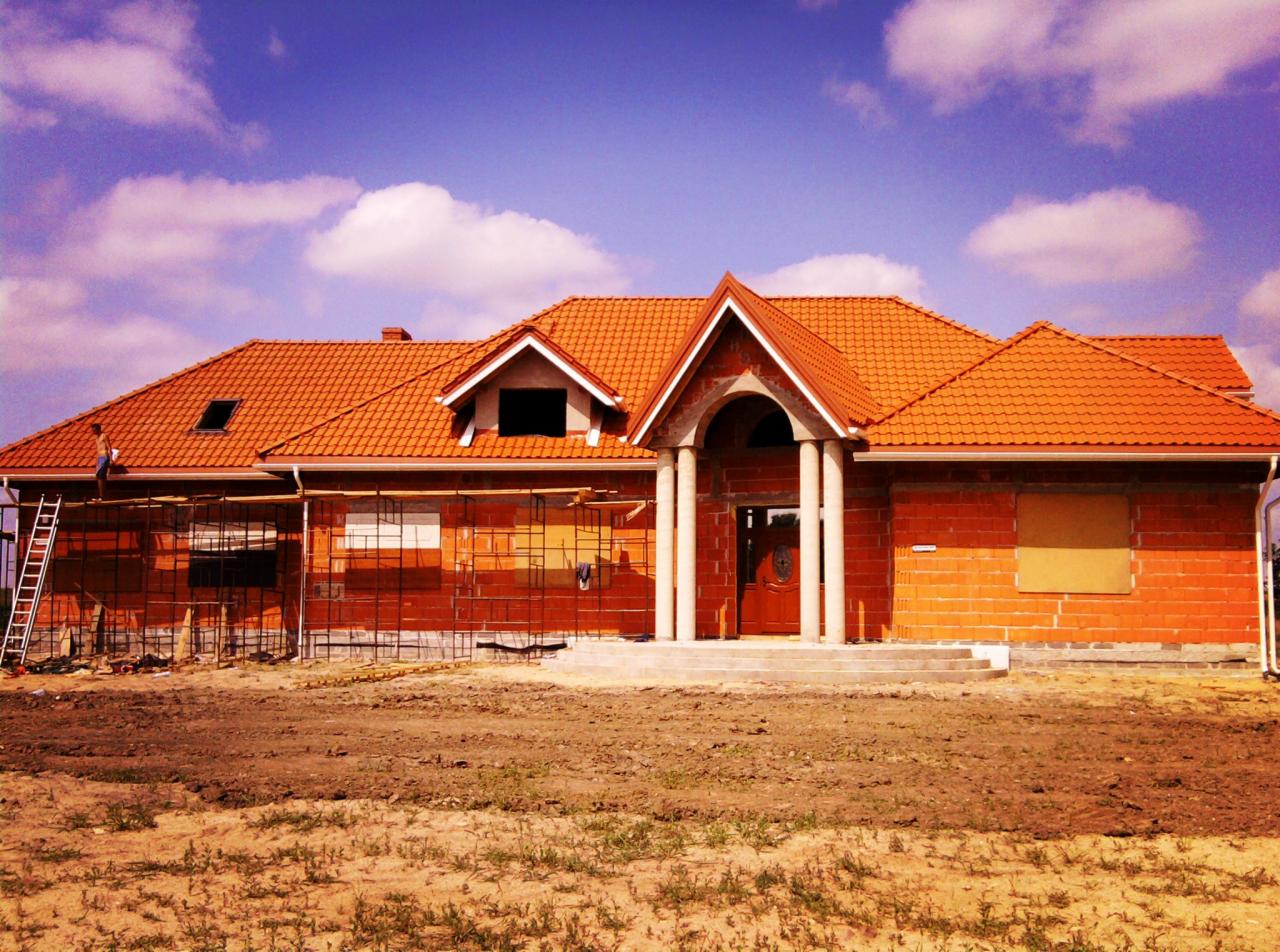 Dom w trakcie budowy z nowym, pomarańczowym dachem z dachówki ceramicznej, widoczne rusztowania i pracownik na dachu, błękitne niebo z chmurami w tle.