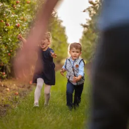 Dwoje dzieci, dziewczynka z uniesioną ręką i chłopiec w kamizelce, biegną w sadzie jabłkowym, z jabłkami leżącymi na trawie, przesłonięci częściowo przez rozmazaną postać w pierwszym planie.