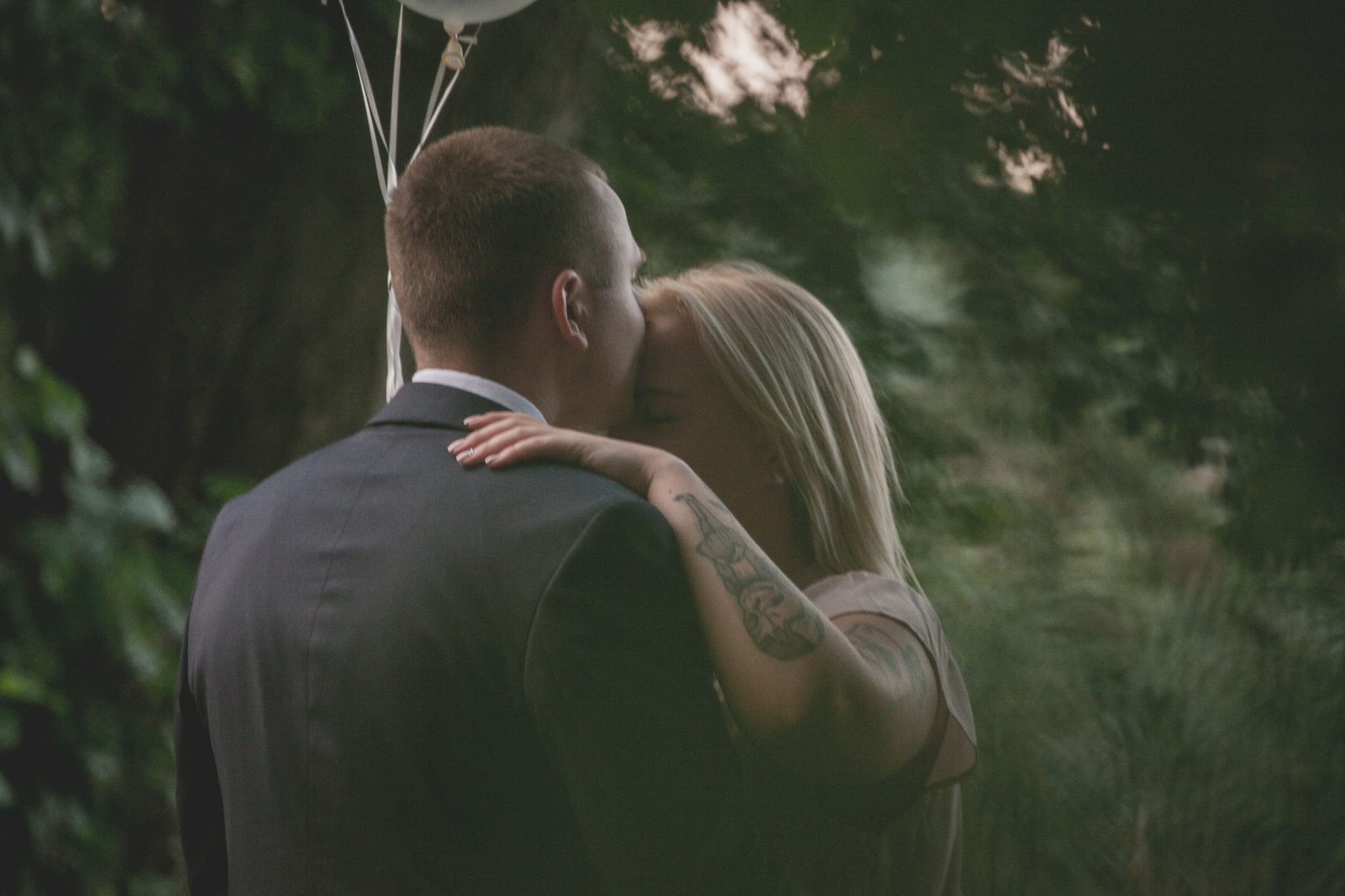 Para w objęciach na tle zieleni, panna młoda z tatuażem na ramieniu opiera dłoń na ramieniu pana młodego w garniturze, nad nimi białe balony.