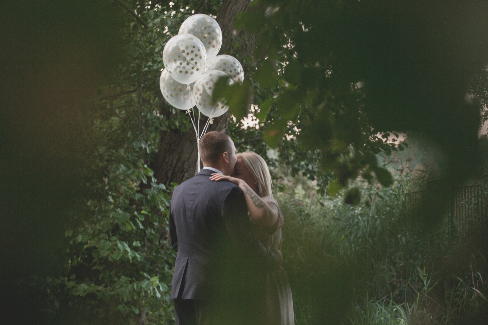 Para obejmująca się w plenerze, mężczyzna w garniturze, kobieta z tatuażem na ramieniu, trzymająca balony w kropki, otoczeni zielenią.