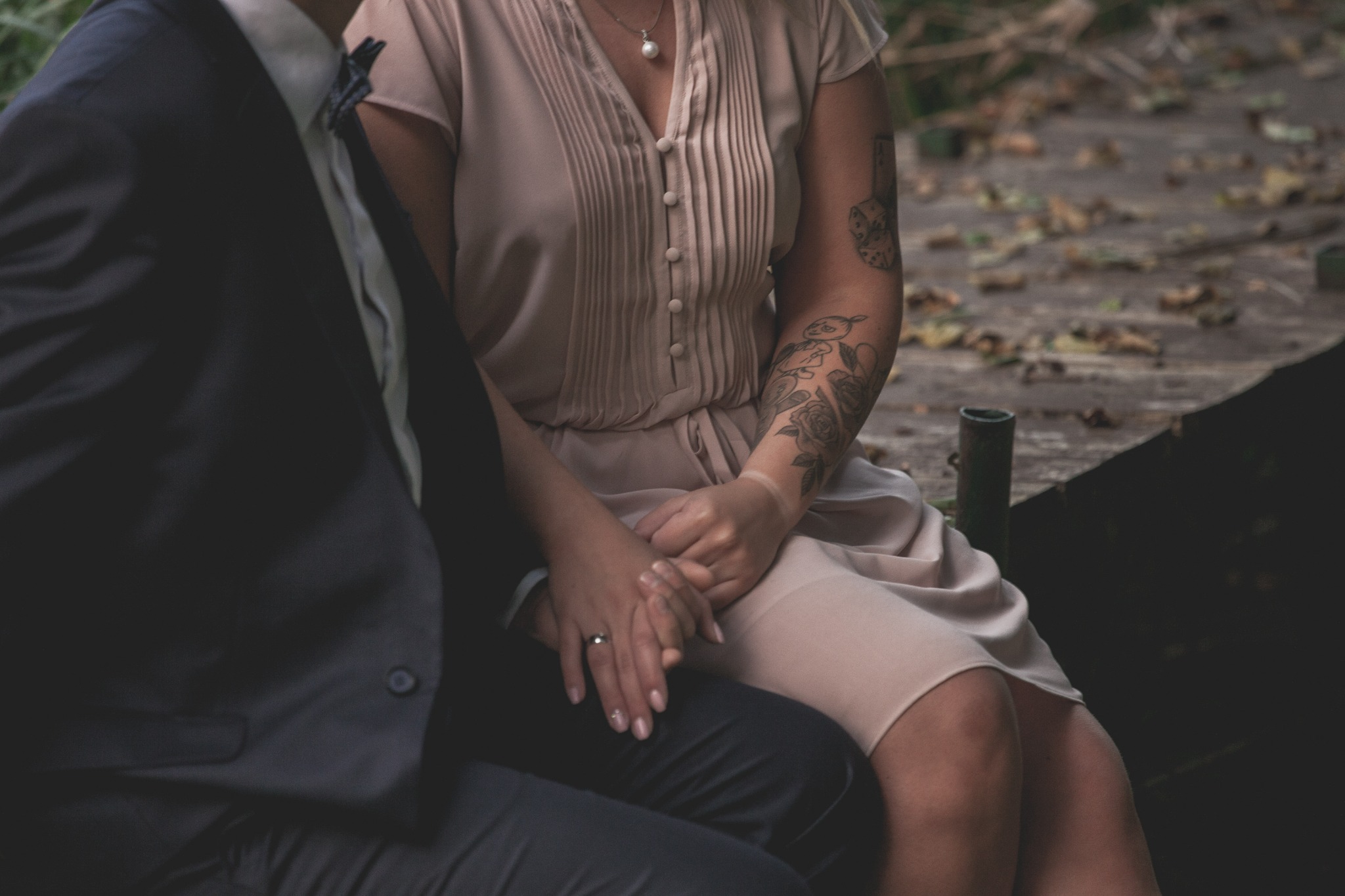 Para trzymająca się za ręce; ona w pudrowej sukience z tatuażami na ramieniu, on w granatowym garniturze z muszką na tle drewnianej podłogi.