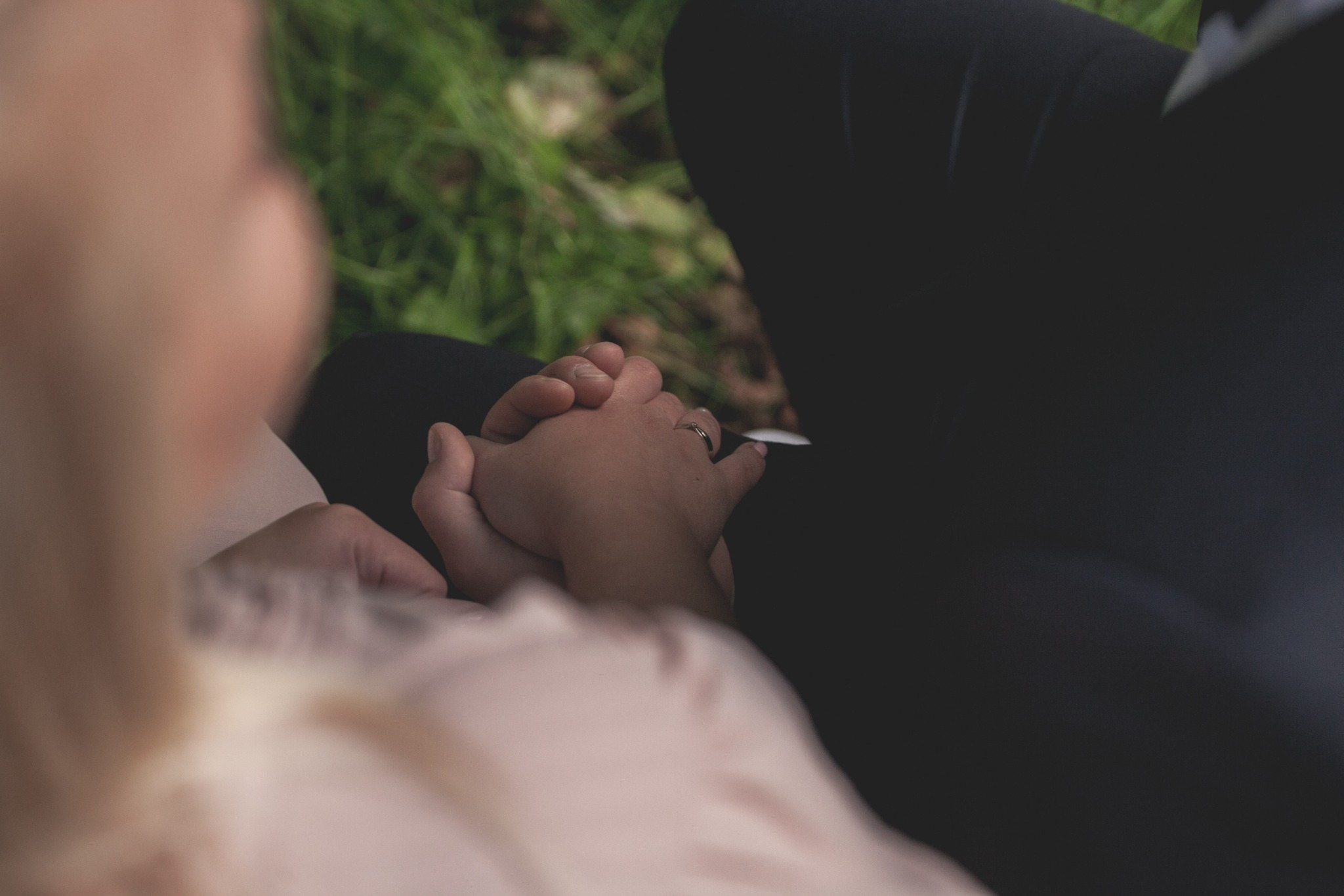 Dłonie pary trzymające się na tle zielonej trawy, widoczny pierścionek zaręczynowy.