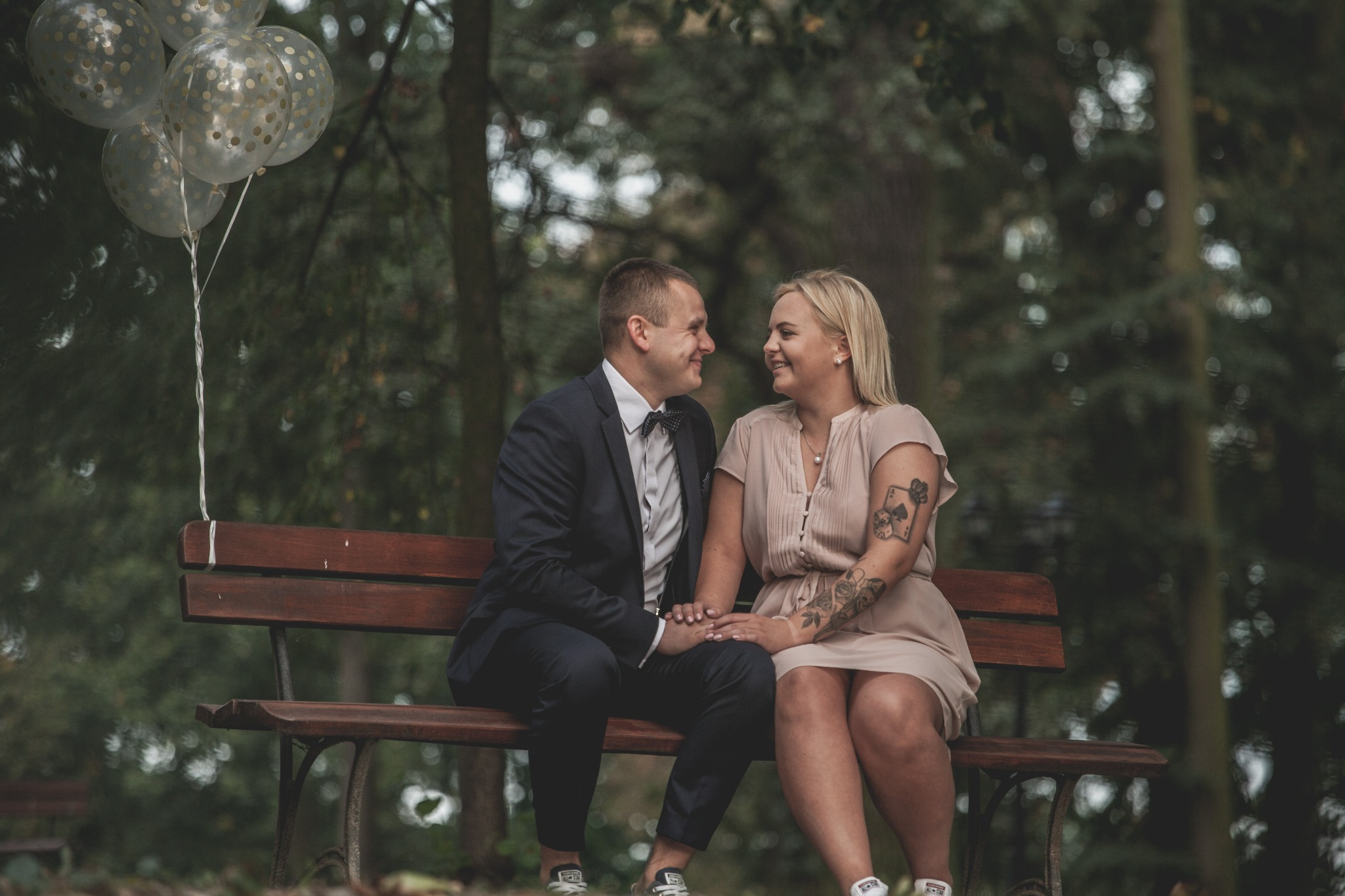 Zakochana para siedzi na ławce w Buku, trzymając się za ręce i patrząc na siebie z czułością. Mężczyzna w garniturze z muchą, kobieta w sukience w kolorze pudrowego różu z tatuażami na ramieniu.