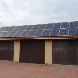 Instalacja fotowoltaiczna na dachu garażu z trzema bramami, widok z zewnątrz na tle pochmurnego nieba.