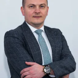 Portret mężczyzny w garniturze w kratę i krawacie w zielono-białą kratkę, z zegarkiem na ręku, na białym tle.