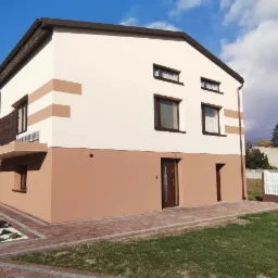 Dwukondygnacyjny dom z jasną elewacją, brązowym pasem dolnym i poziomymi akcentami, widok z poziomu gruntu, z kostką brukową i małym ogródkiem.