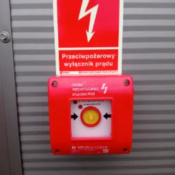 Czerwony, natynkowy przycisk przeciwpożarowy wyłącznika prądu z żółtym guzikiem, umieszczony na szarej, metalowej ścianie z poziomymi przetłoczeniami, widoczna tabliczka informacyjna z symbolem...