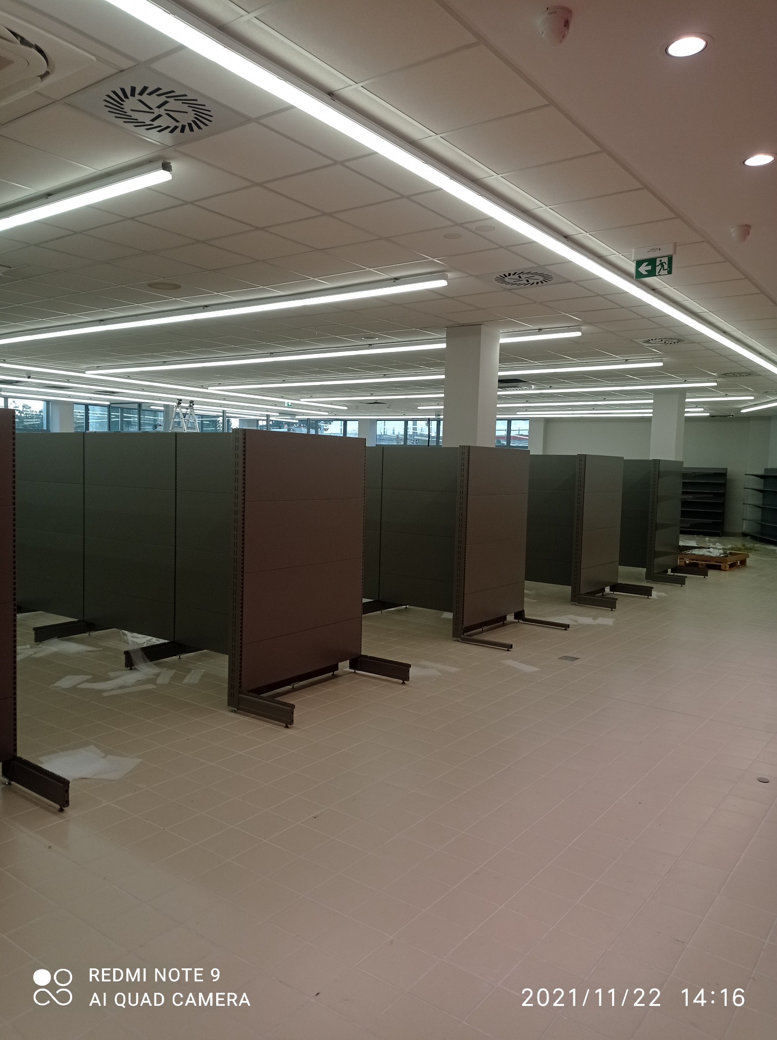 Montaż regałów sklepowych w nowym, przestronnym lokalu z jasnym oświetleniem i widocznymi elementami wentylacji na suficie.