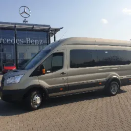 Beżowy bus Ford Transit z przyciemnianymi szybami, zaparkowany na tle salonu Mercedes-Benz w słoneczny dzień.