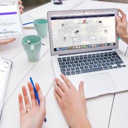 LogMap - Osoby korzystające z urządzeń (laptop, tablet, smartfon) z otwartymi stronami internetowymi kalkulatorów transportowych i platform logistycznych na białym stole, w tle dwie zielone filiżanki.