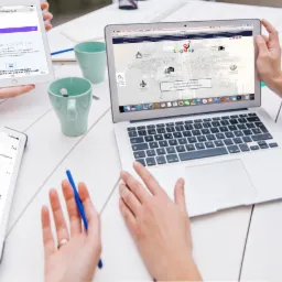 Osoby korzystające z urządzeń (laptop, tablet, smartfon) z otwartymi stronami internetowymi kalkulatorów transportowych i platform logistycznych na białym stole, w tle dwie zielone filiżanki.