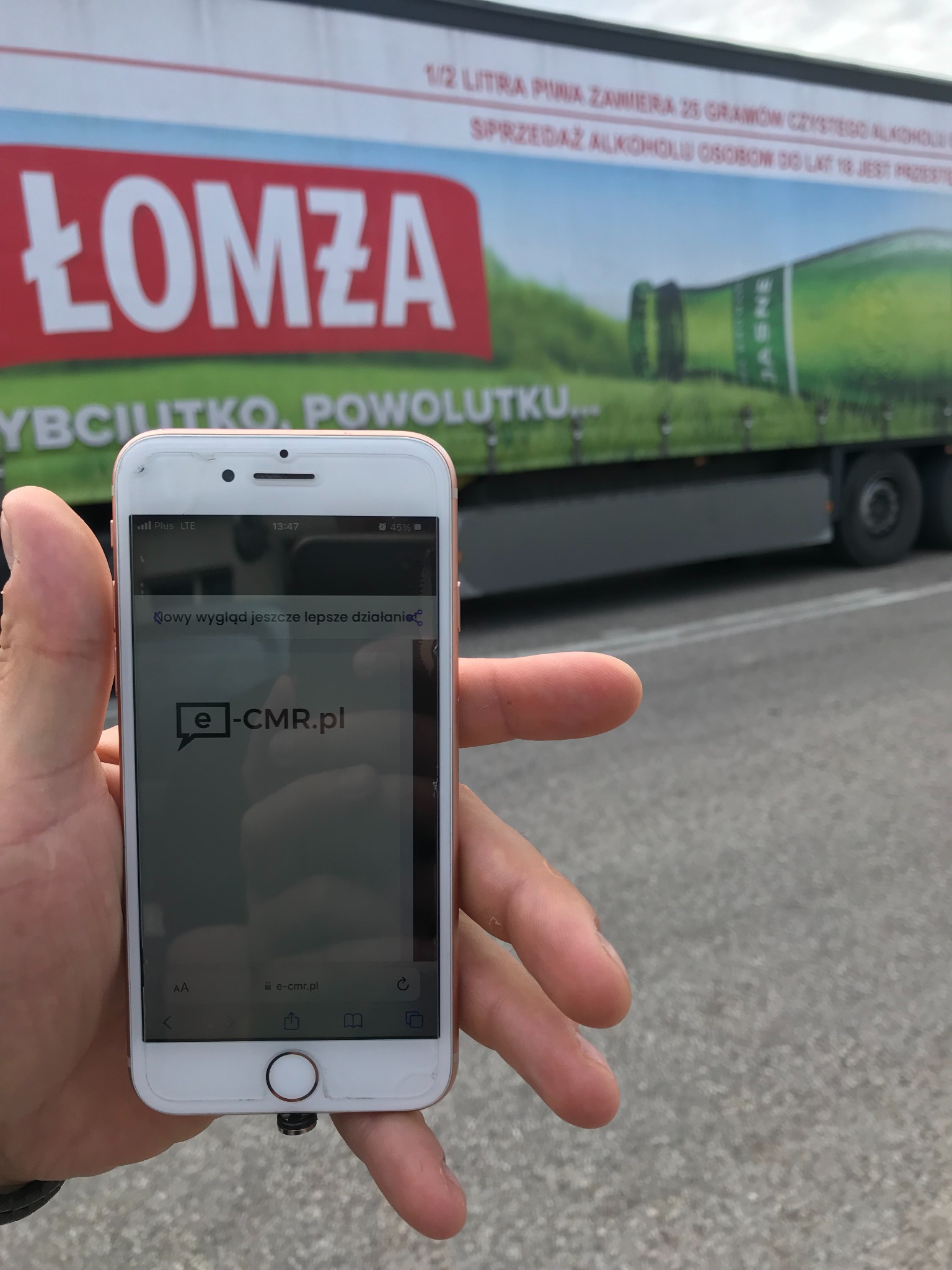 Smartfon wyświetlający stronę e-cmr.pl trzymany w dłoni, na drugim planie naczepa ciężarówki z reklamą piwa Łomża.