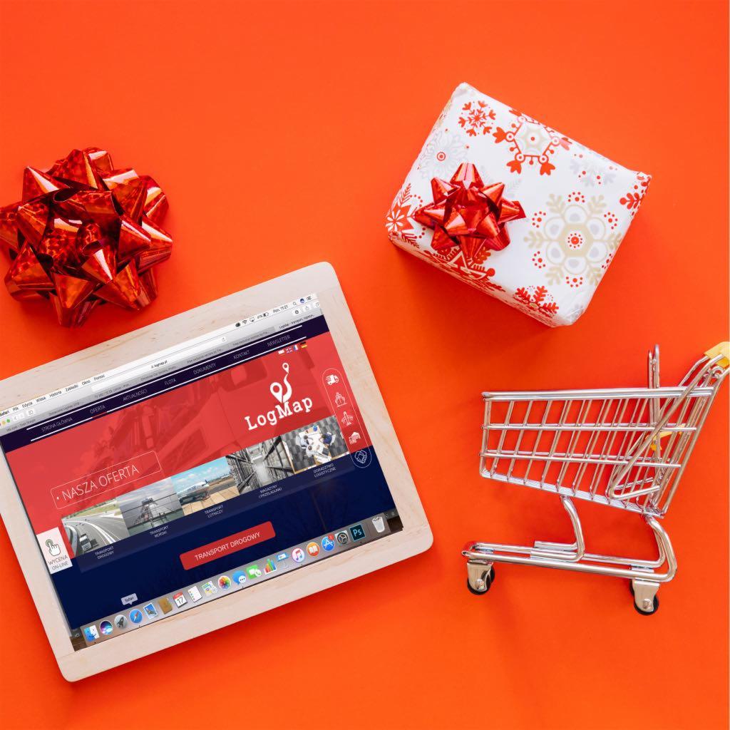 Tablet wyświetlający stronę LogMap z ofertą transportu drogowego, obok prezent ze wstążką i miniaturowy wózek sklepowy na pomarańczowym tle.